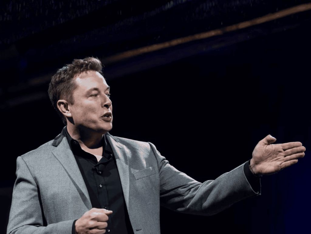 Male presenter in a gray blazer gesturing with his hands on stage during a talk.