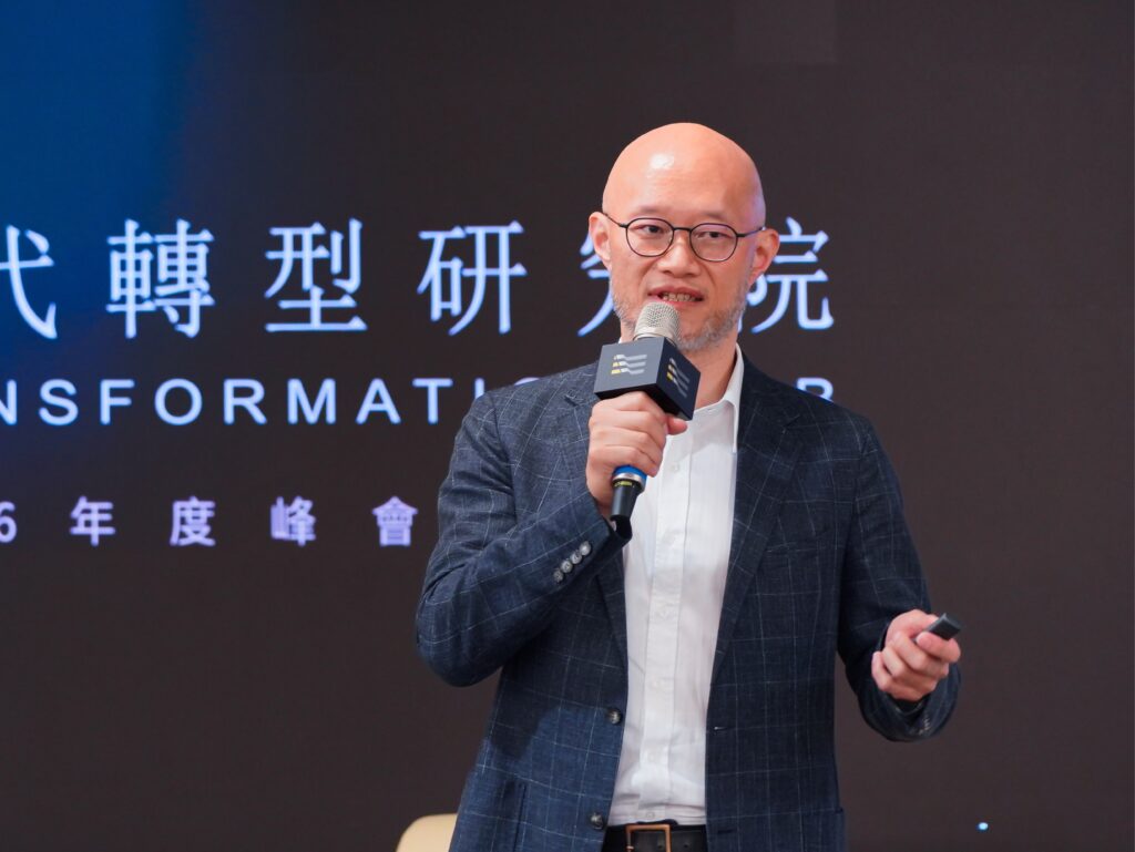 Male speaker with glasses and a shaved head holding a microphone on stage, with large Chinese characters projected on a dark backdrop behind him.