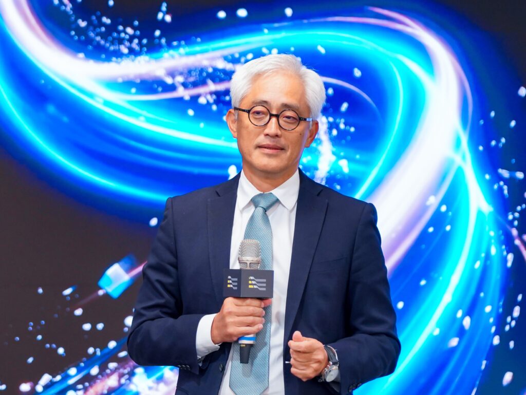 Male presenter in a navy suit and light blue tie speaks into a handheld microphone on a stage with blue neon swirls.