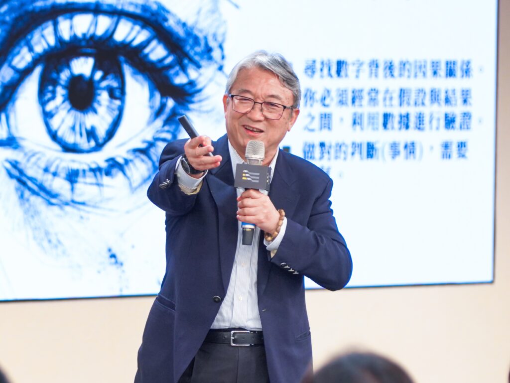 Man in a navy suit speaking into a handheld microphone on stage, gesturing with his hand, with a large blue eye image on a screen behind him.