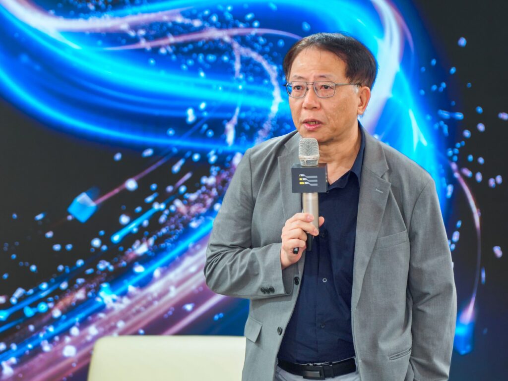 Man in a gray suit speaks into a microphone during a presentation, with a blue abstract light background behind him.