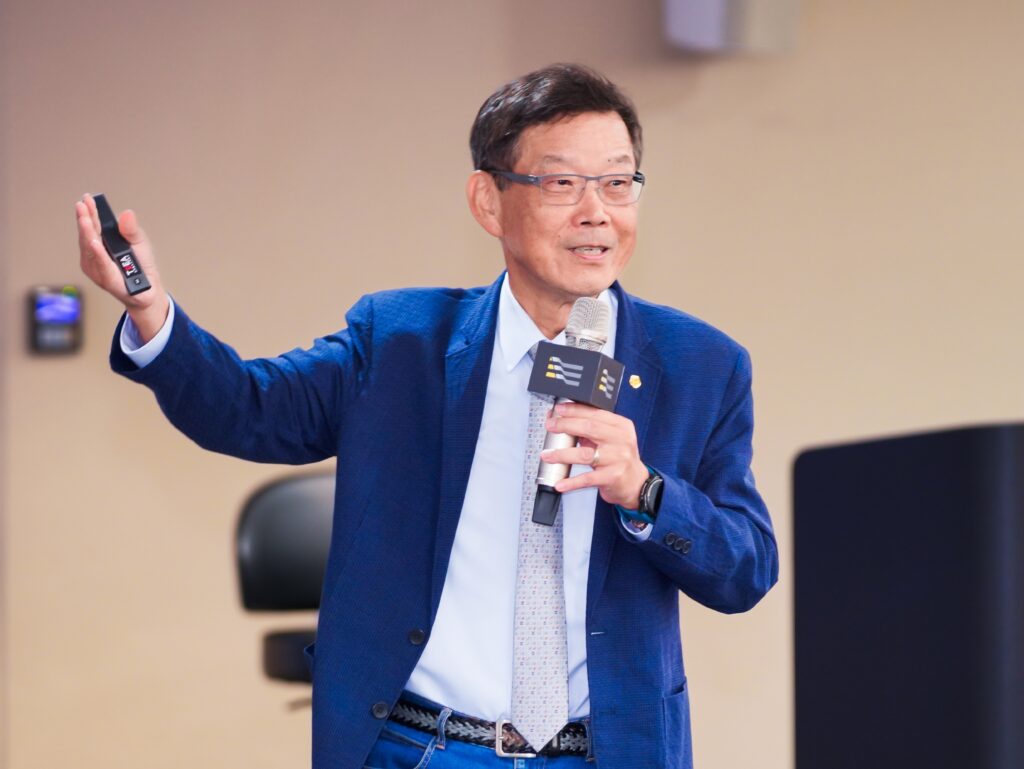 Mid-speech: man in a blue blazer and glasses holds a microphone and a remote, gesturing with his right hand in a conference setting.