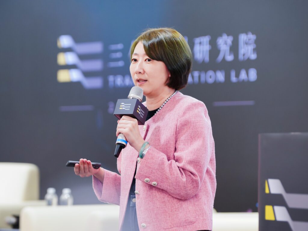 Woman in a pink blazer speaks into a microphone on stage, holding a remote in one hand with a blurred conference backdrop behind her.