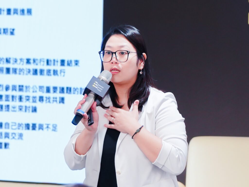 Woman in a white blazer and black top speaks into a microphone during a presentation, with a blue-screened backdrop of Chinese text behind her.