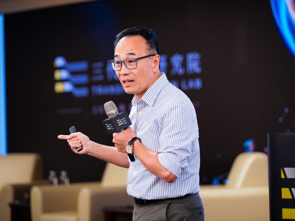Man with glasses and striped shirt speaks into a handheld microphone during a panel discussion on a stage.