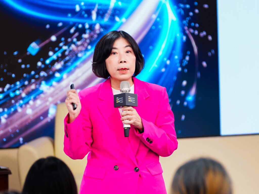 Female presenter in a bright pink suit speaks into a microphone on stage against a blue abstract background.