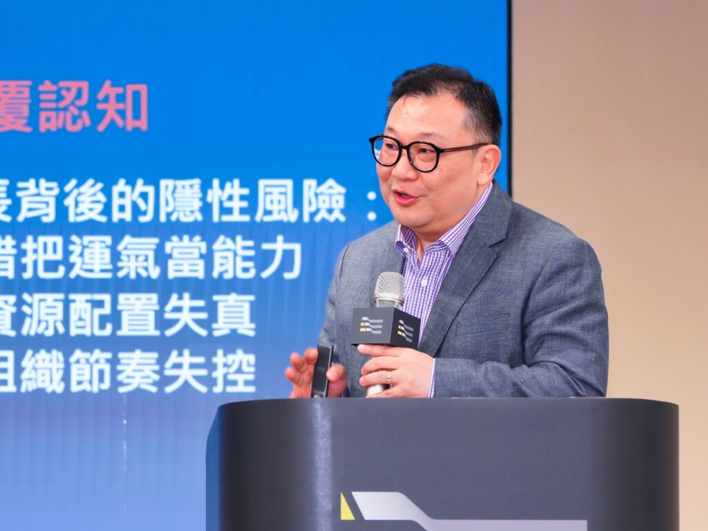 Man with glasses in a gray suit speaks at a podium, blue backdrop with Chinese text behind him.