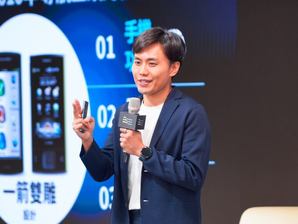Male presenter in a navy blazer holding a microphone, speaking on stage with a large tech backdrop displaying smartphones.
