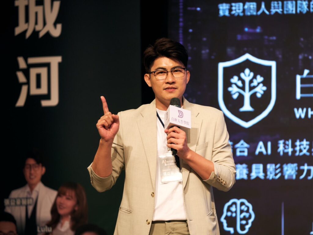 Presenter in a light suit speaks into a handheld microphone on stage, raising one finger as he talks, with a dark backdrop and blue logo graphics behind him.