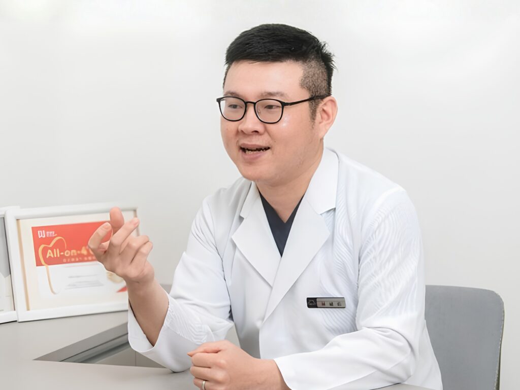 Medical professional wearing a white coat and glasses explains something, gesturing with his hands at a desk with a red poster nearby.