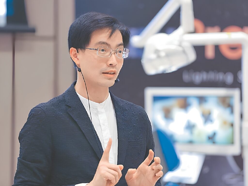 Asian man in a dark blazer with a headset mic, gesturing as he speaks at a presentation. Spotlit background blurred behind him.