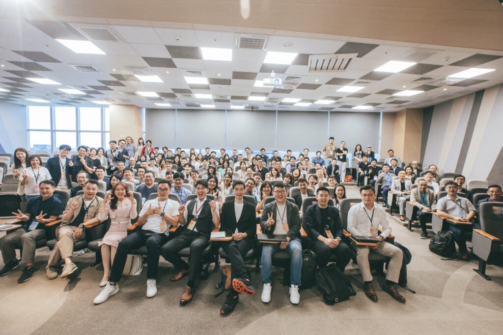 Large group of conference attendees posing for a group photo in an auditorium, many smiling faces and seated with lanyards visible