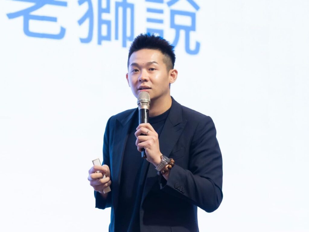 Male presenter in a dark suit speaks into a handheld microphone on stage, with a beaded bracelet visible on his wrist.