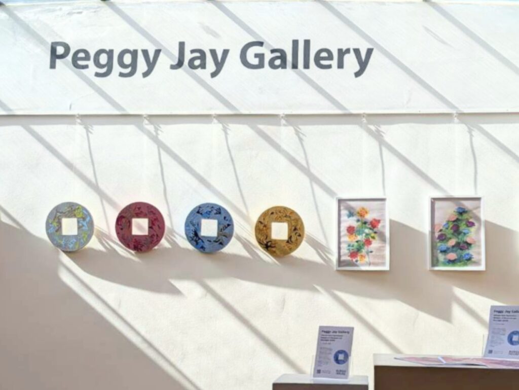 signage reading 'Peggy Jay Gallery' with circular colorful wall art pieces and framed floral prints beneath the letters