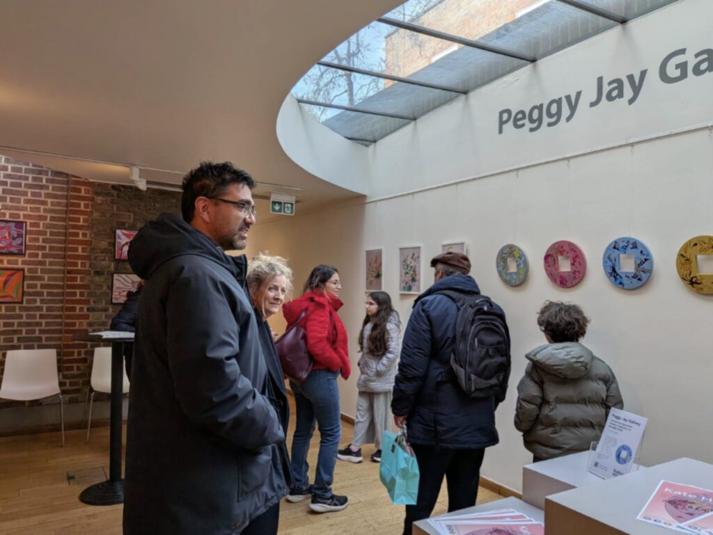 Group of visitors in jackets looking at art on white walls inside an exhibition space, Peggy Jay Gallery sign visible above.