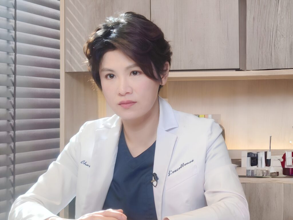 Woman in a white lab coat and navy scrubs seated at a desk, with wooden shelves behind her.