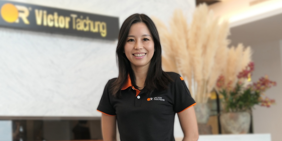Smiling woman in a black polo with orange trim standing in a reception area with a Victor Taichung sign in the background.