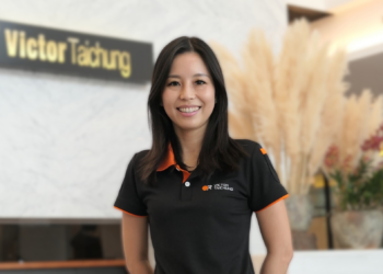 Smiling woman in a black polo with orange trim standing in a reception area with a Victor Taichung sign in the background.